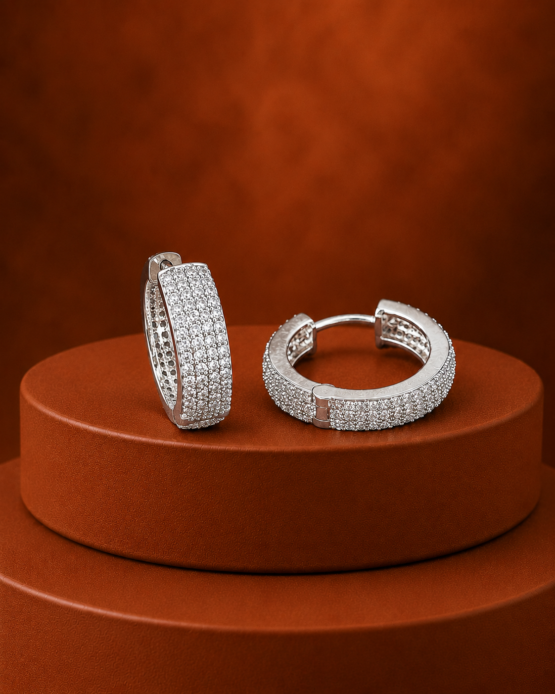 A pair of silver huggie hoop earrings paved with small clear stones, displayed on a round terracotta pedestal against a matching textured brown background.
