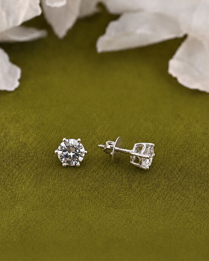 A pair of silver stud earrings with clear round-cut gemstones, one shown from the front and the other from the side/back. They are displayed on a textured olive green surface with soft white flower petals in the background.