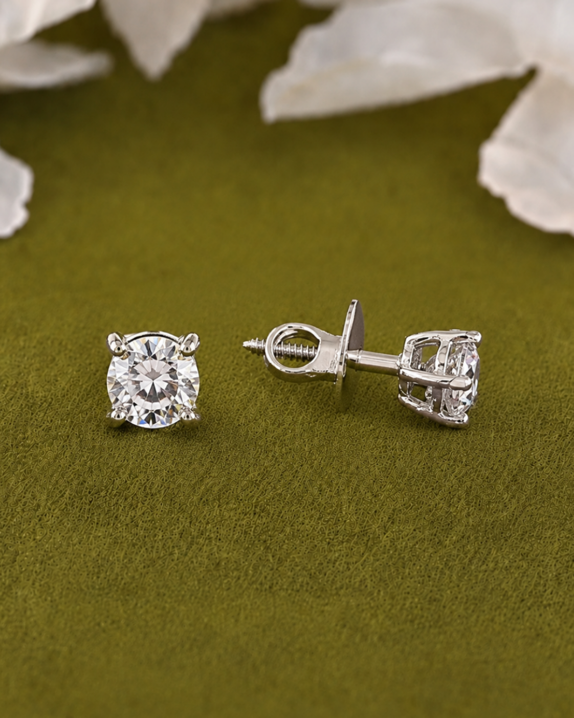 A pair of silver stud earrings with round-cut clear gemstones, one shown from the front and the other from the side/back. They are displayed on a textured olive green surface with soft white flower petals in the background.