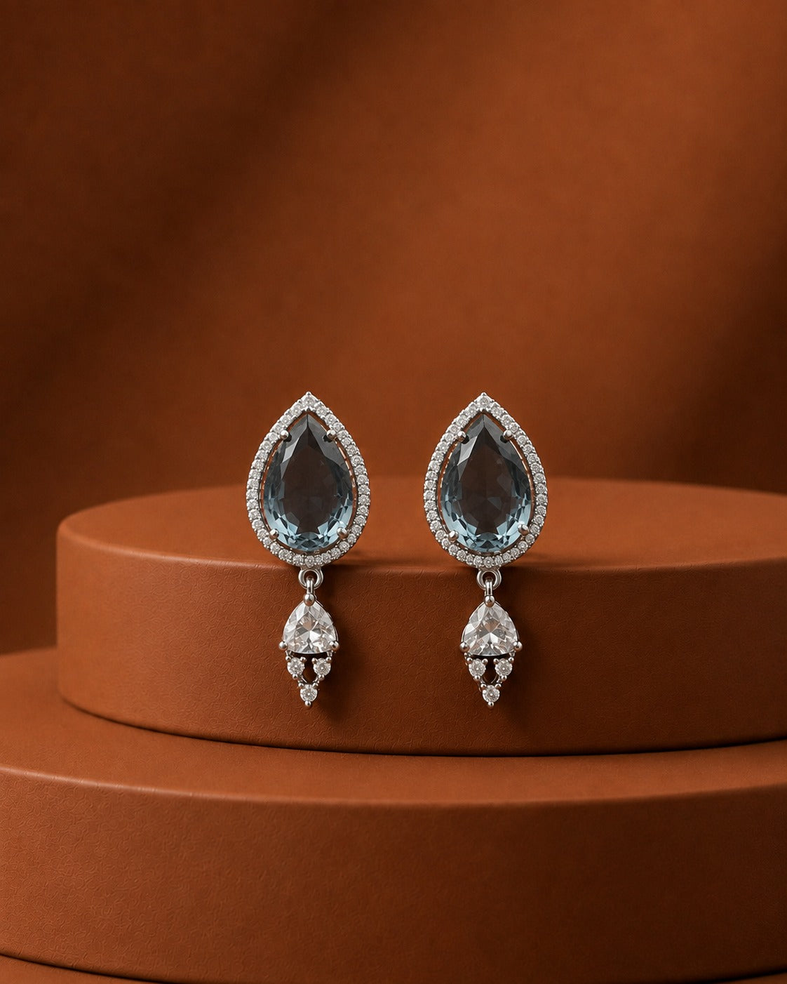 A pair of silver teardrop dangle earrings with light blue gemstone centers and clear accent stones, displayed on a round brown pedestal against a warm brown background.