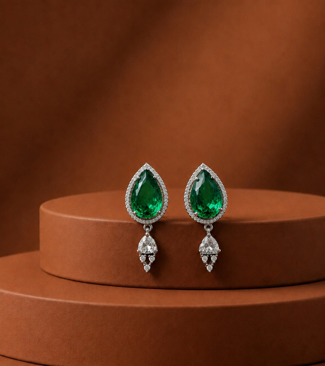 A pair of silver teardrop dangle earrings with vibrant green gemstone centers and clear accent stones, displayed on a brown cylindrical pedestal against a matching brown background.