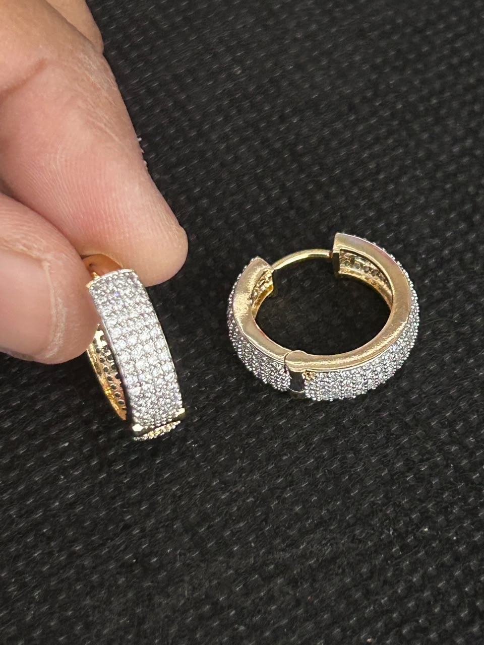 A pair of small gold huggie hoop earrings covered in clear pavé stones, shown with a person's fingers holding one earring on a black textured background.