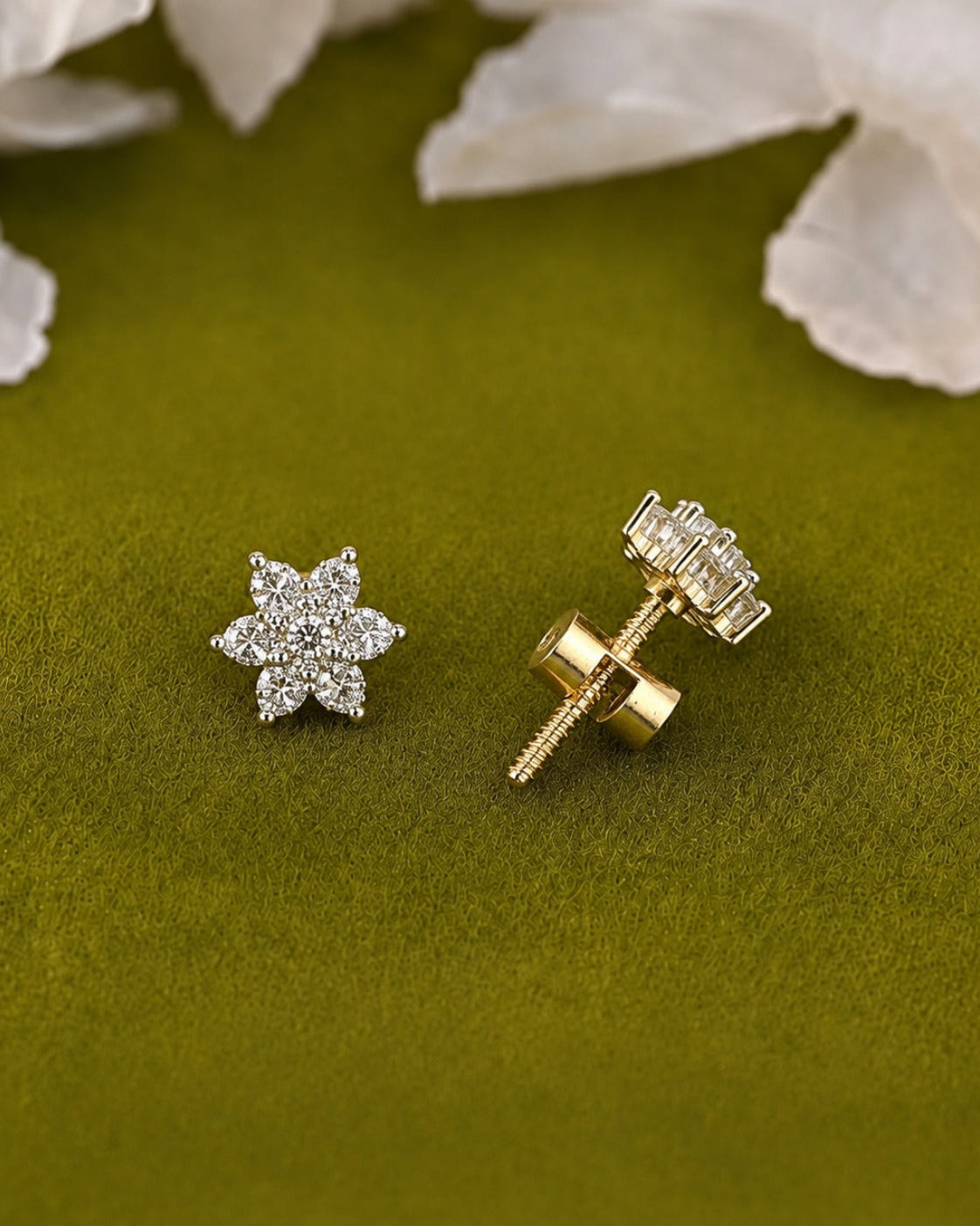 A pair of small stud earrings on an olive green textured surface with blurred white flowers in the background. One earring is shown front-facing as a silver-toned flower cluster, and the other is turned sideways showing the gold-toned screw-back post and clear stones.