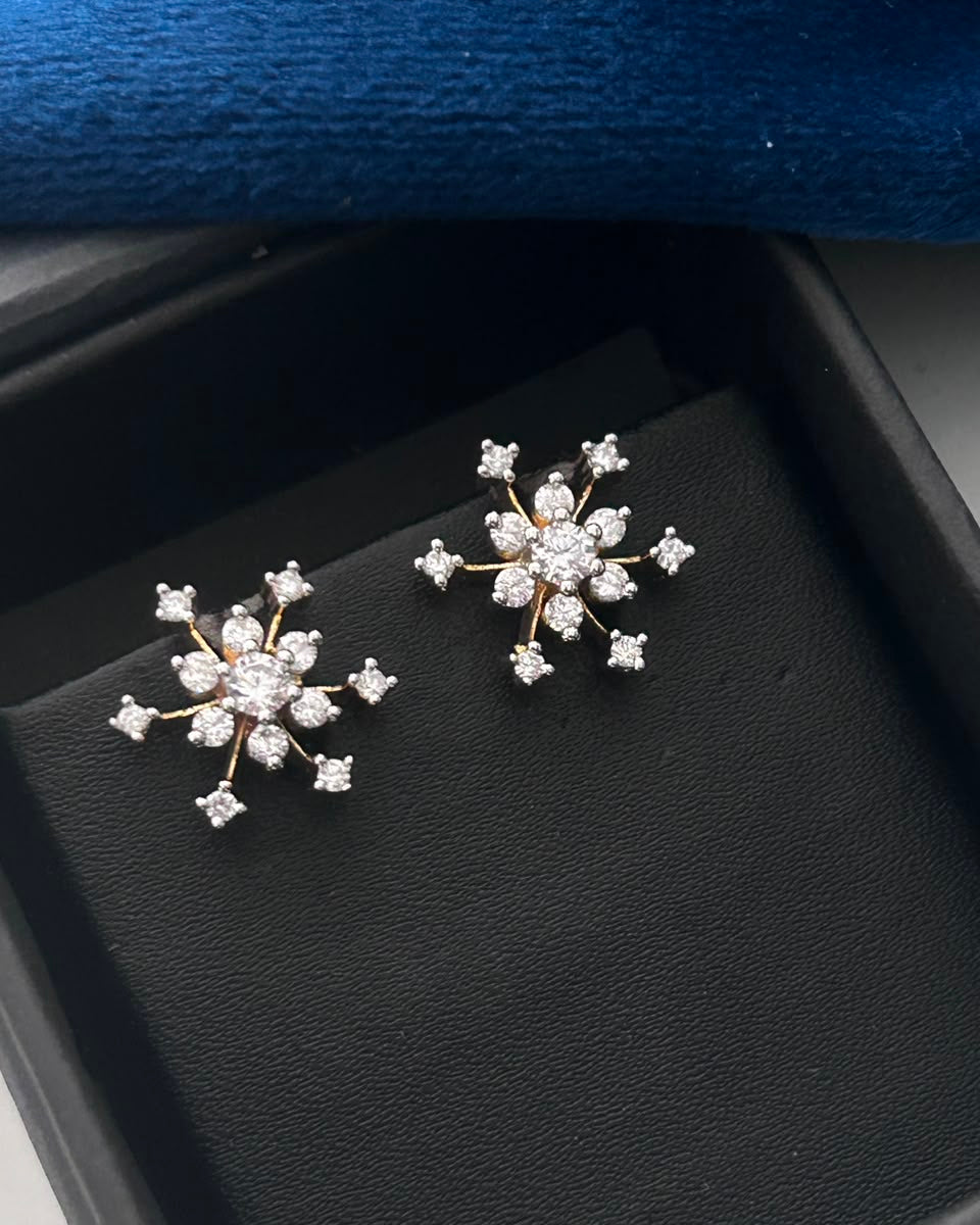 A pair of snowflake-shaped stud earrings with clear gemstones and gold-toned settings, displayed on a black jewelry box insert with a blue velvet lid edge visible above.