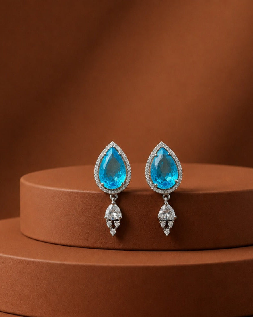A pair of teardrop-shaped drop earrings with bright blue gemstones surrounded by small clear stones, displayed on a round brown pedestal against a warm brown background.