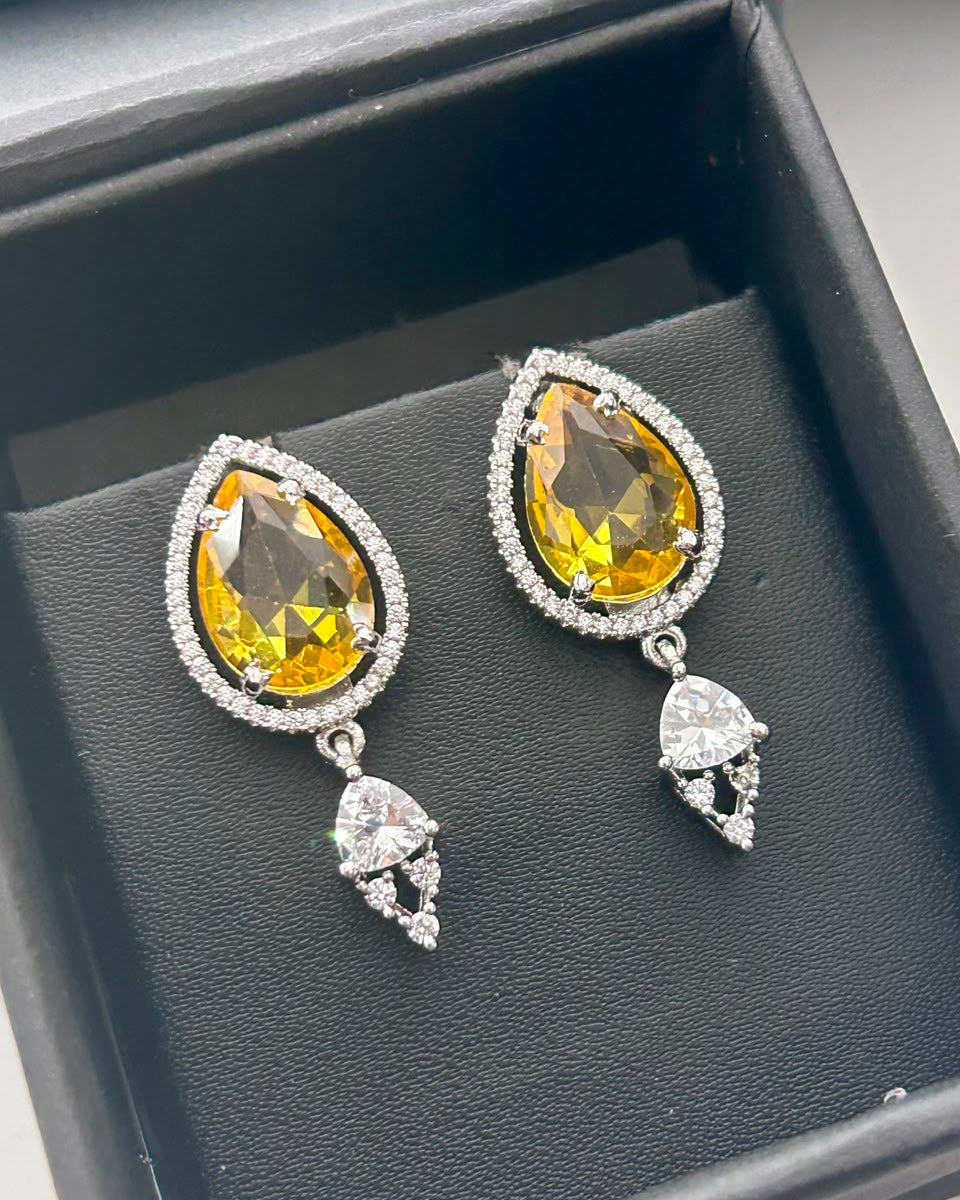A pair of teardrop-shaped drop earrings displayed in a black jewelry box. Each earring features a large faceted yellow gemstone surrounded by a halo of small clear stones, with a dangling clear teardrop-shaped stone below.