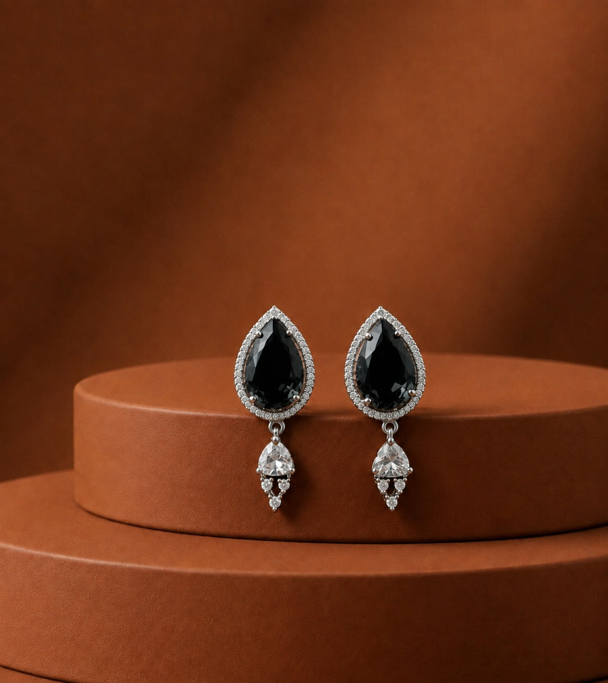 A pair of teardrop-shaped drop earrings with black teardrop gemstones surrounded by small clear stones, hanging from silver-toned settings, displayed on a round brown pedestal against a warm brown background.