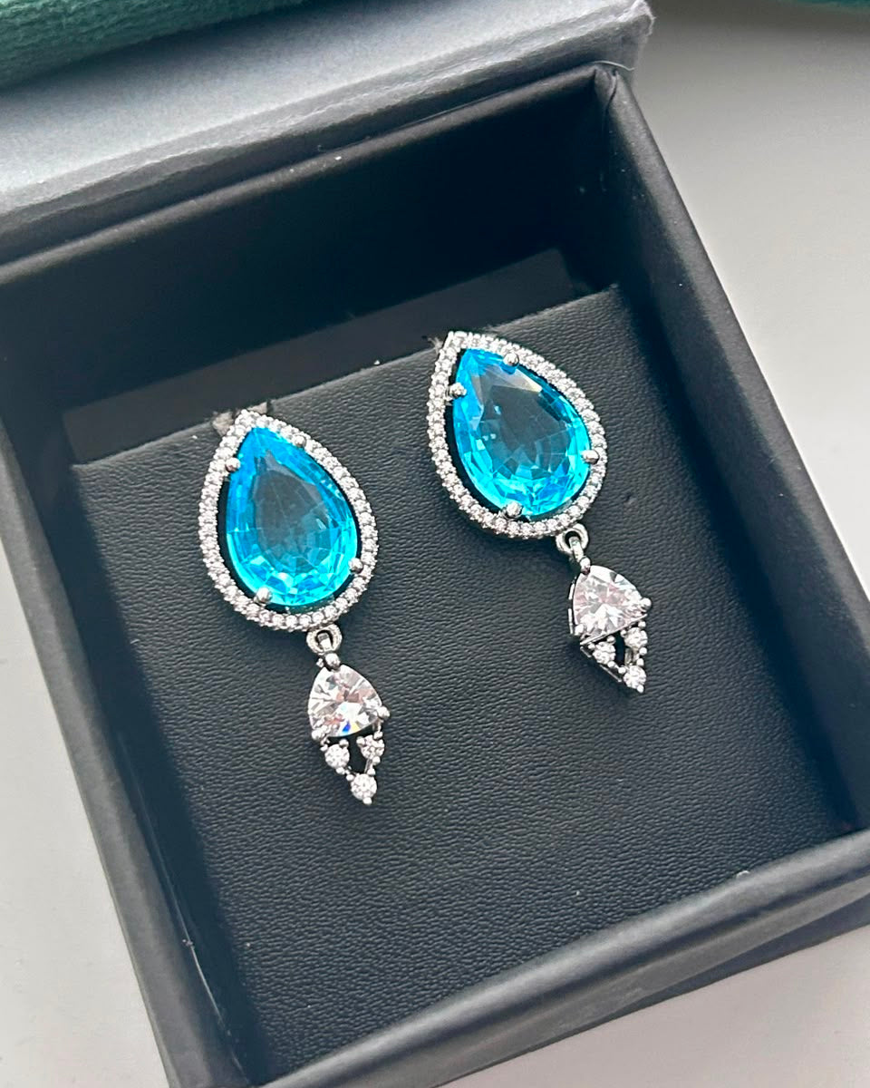 A pair of teardrop-shaped drop earrings with bright blue gemstones and clear rhinestone accents, displayed inside a black jewelry box.