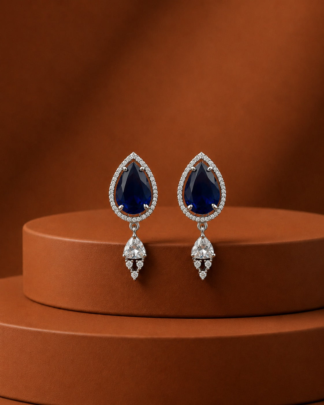 A pair of teardrop-shaped drop earrings with deep blue gemstones surrounded by small clear stones, hanging from silver-toned settings, displayed on a round brown pedestal against a warm brown background.
