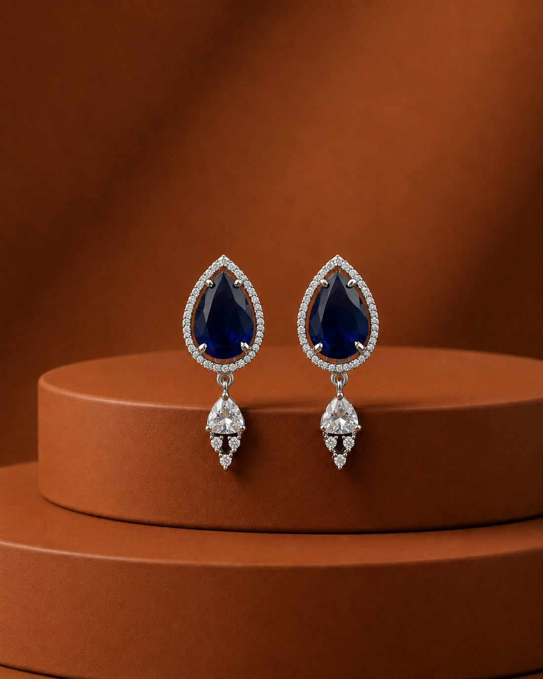 A pair of teardrop-shaped drop earrings with deep blue gemstones surrounded by small clear stones, hanging from silver-toned settings, displayed on a round brown pedestal against a warm brown background.