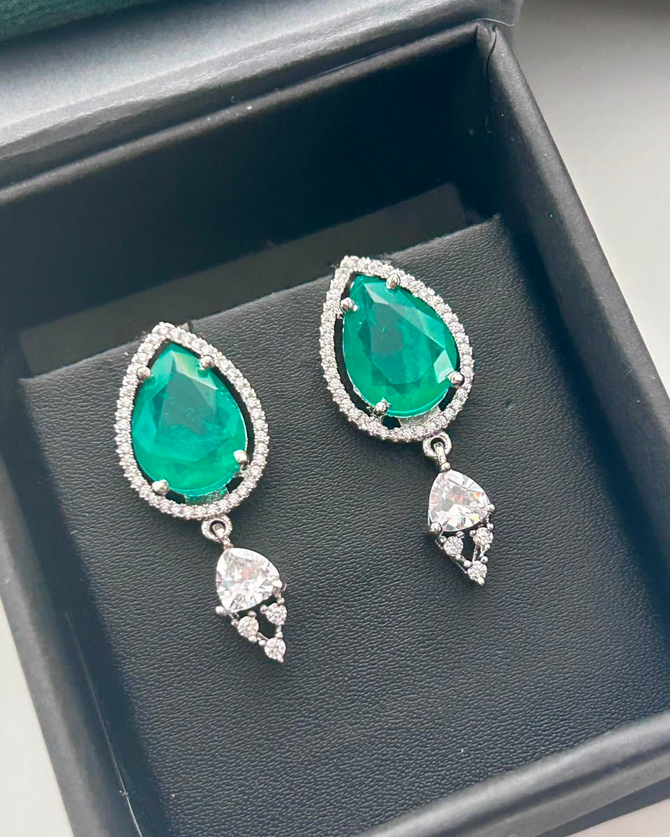 A pair of teardrop-shaped drop earrings with large green gemstones surrounded by small clear stones, displayed inside a black jewelry box.