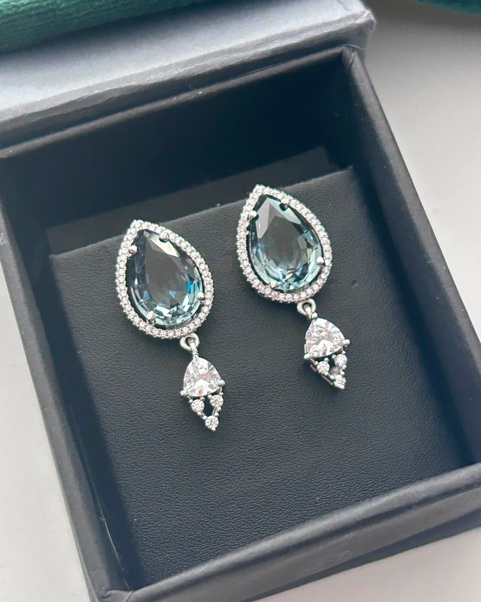 A pair of teardrop-shaped drop earrings with large light blue gemstones, surrounded by small clear stones and hanging from silver-toned settings, displayed inside an open dark jewelry box.
