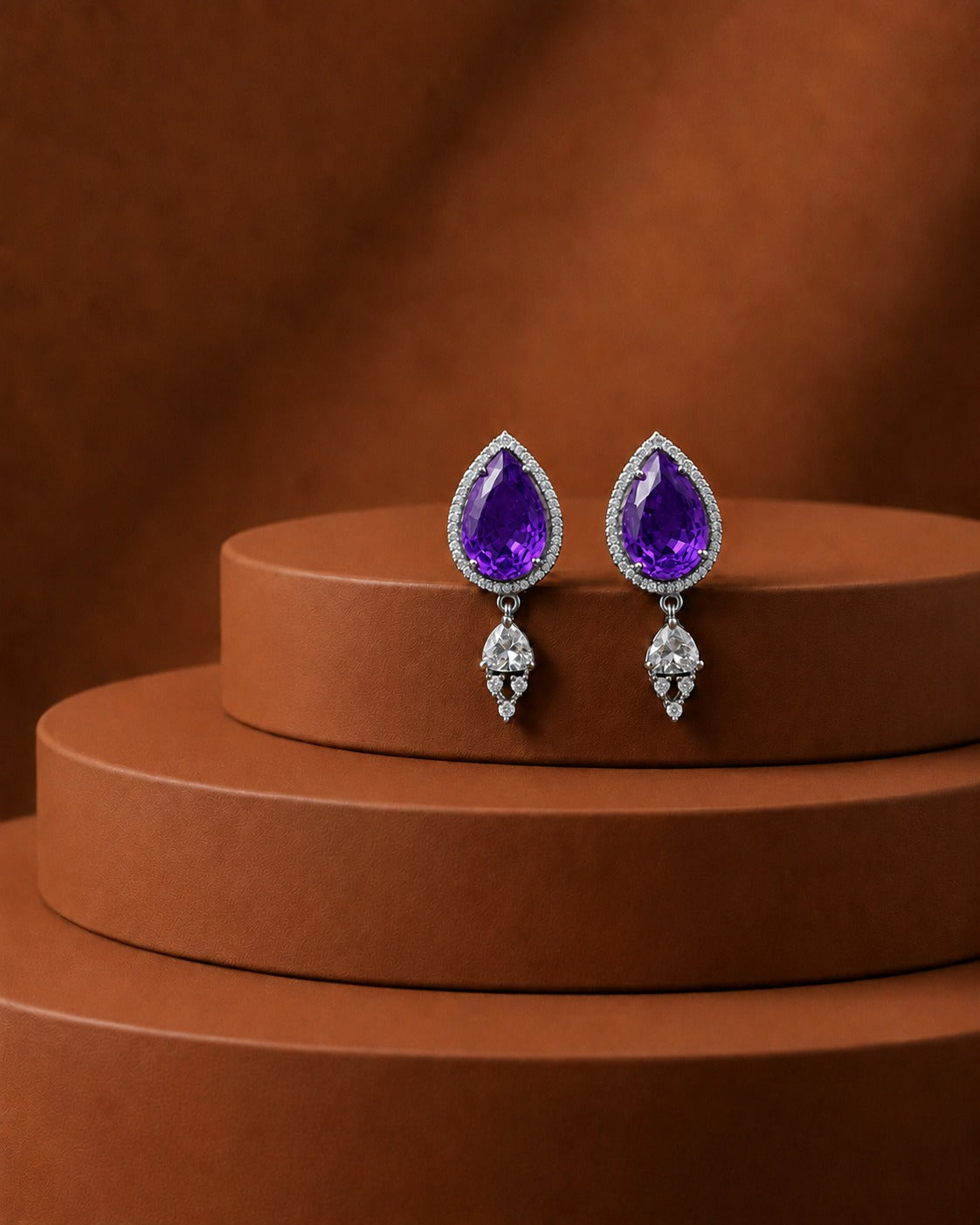 A pair of teardrop-shaped drop earrings with large purple gemstones and small clear stones, displayed on a brown tiered pedestal against a matching brown background.