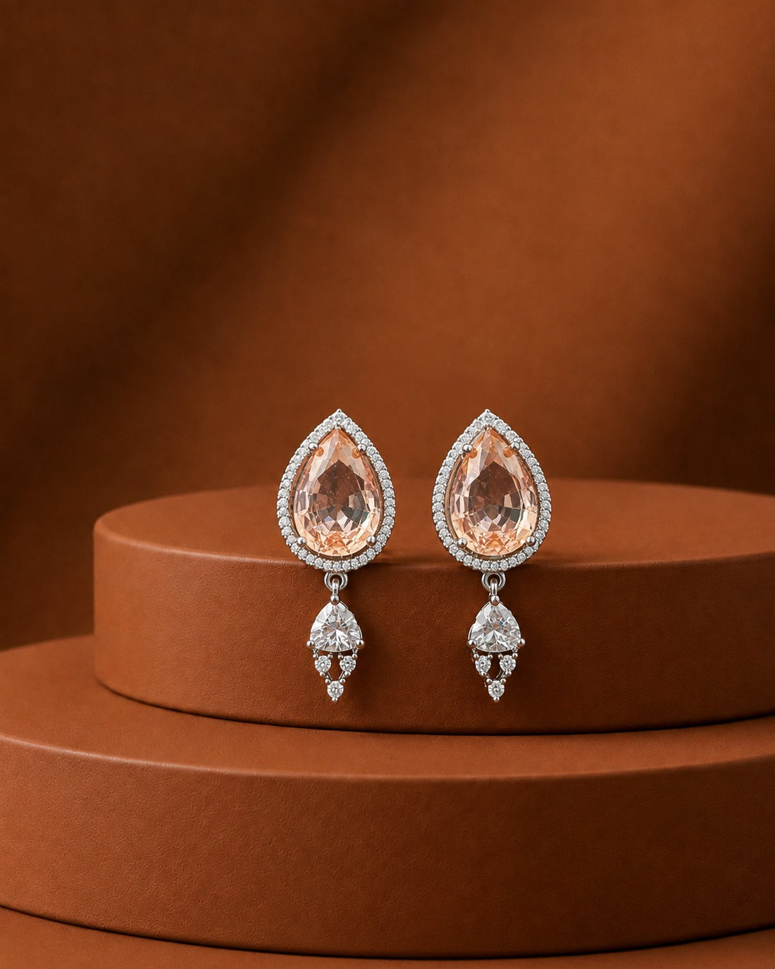 A pair of teardrop-shaped drop earrings with peach-colored center stones surrounded by small clear stones, hanging from silver-toned settings, displayed on a round brown pedestal against a matching brown background.