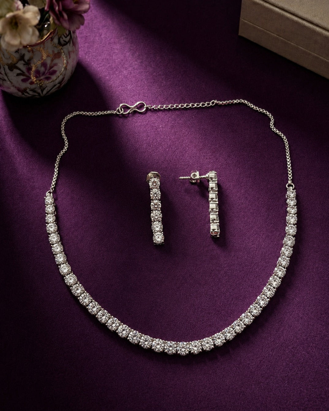 A silver-tone necklace and matching drop earrings set with clear stones, arranged on a rich purple fabric background with a decorative vase and box partially visible.