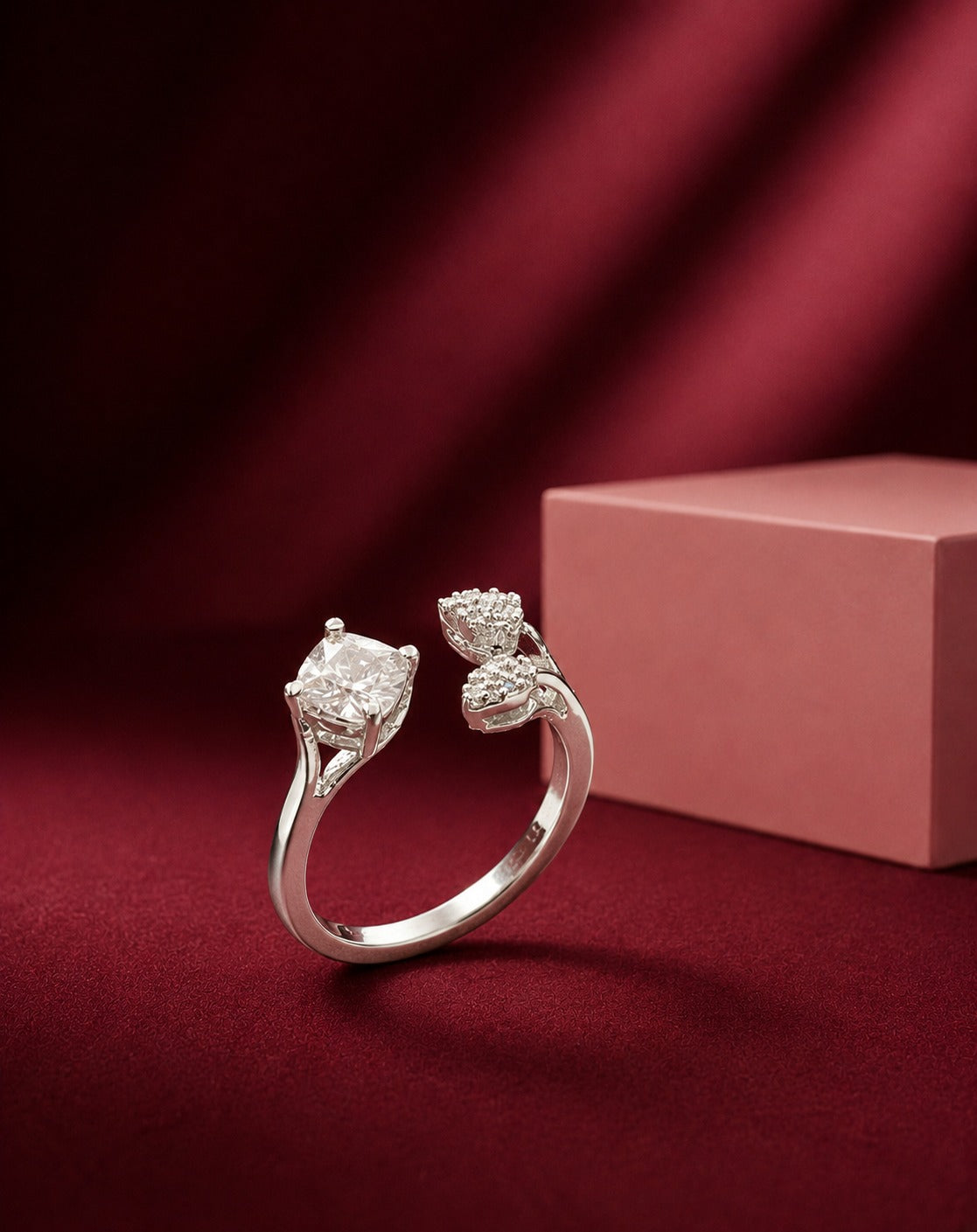A silver-toned diamond ring with a large cushion-cut center stone and smaller diamond accents, displayed on a dark red fabric surface next to a pink jewelry box.