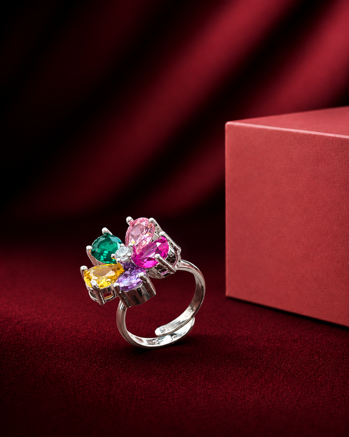 A silver-toned ring with a cluster of colorful gemstones including green, pink, yellow, and purple, displayed next to a red gift box on a dark red velvet background.