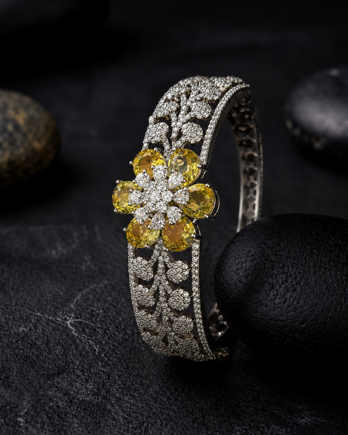 A silver-toned wide cuff bracelet featuring a central flower motif set with yellow gemstones and pavé-set clear stones, displayed on a dark textured surface with black stones.