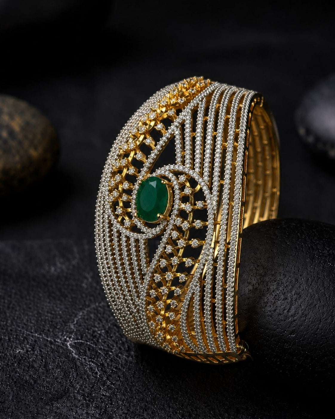 A wide gold and silver bangle bracelet with intricate woven patterns, a central green oval stone, and small clear stones, displayed on a dark textured surface with black stones in the background.