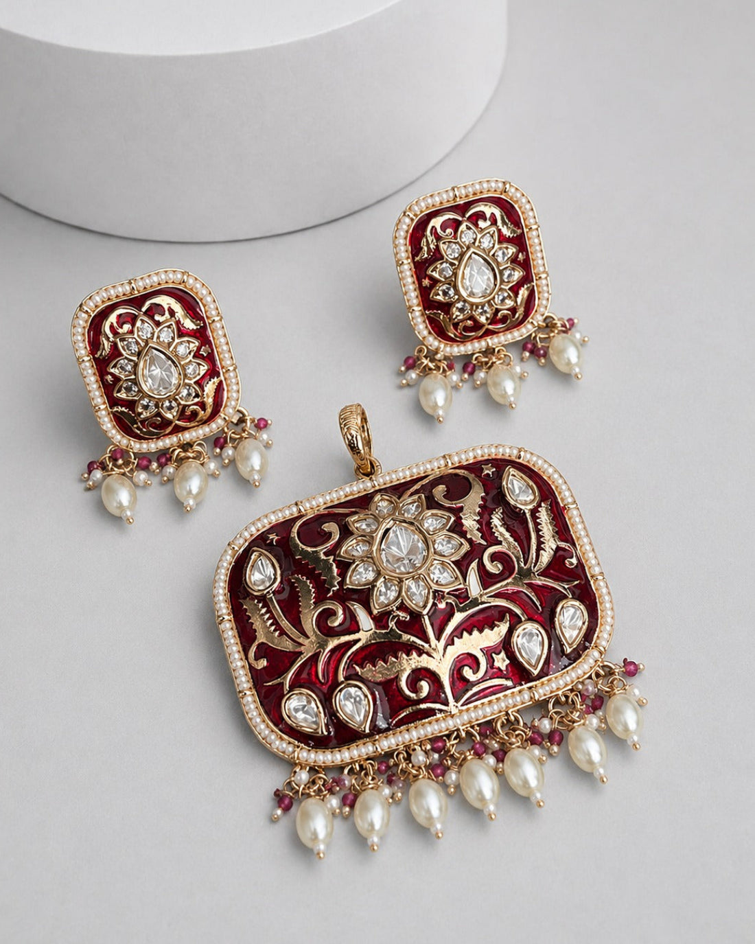 An ornate Indian jhumka earrings and matching pendant set with deep red enamel, gold-plated filigree, and dangling white faux pearls on a light gray background.