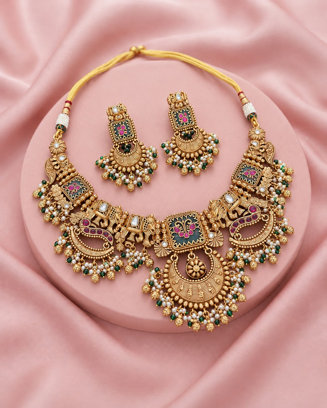 An ornate gold-toned traditional necklace set with matching earrings, decorated with green and pink stone accents and white bead details, displayed on a pink satin background.