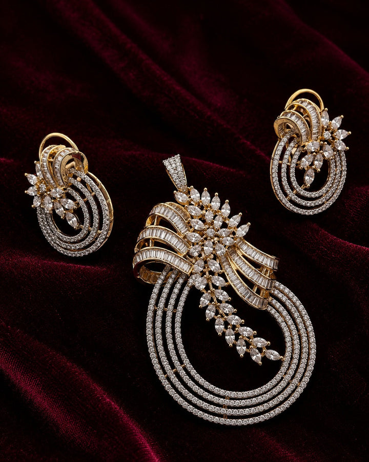 An ornate gold jewelry set featuring a large pendant, matching earrings, and a slim bracelet, all adorned with sparkling clear stones and set against a dark red velvet background.