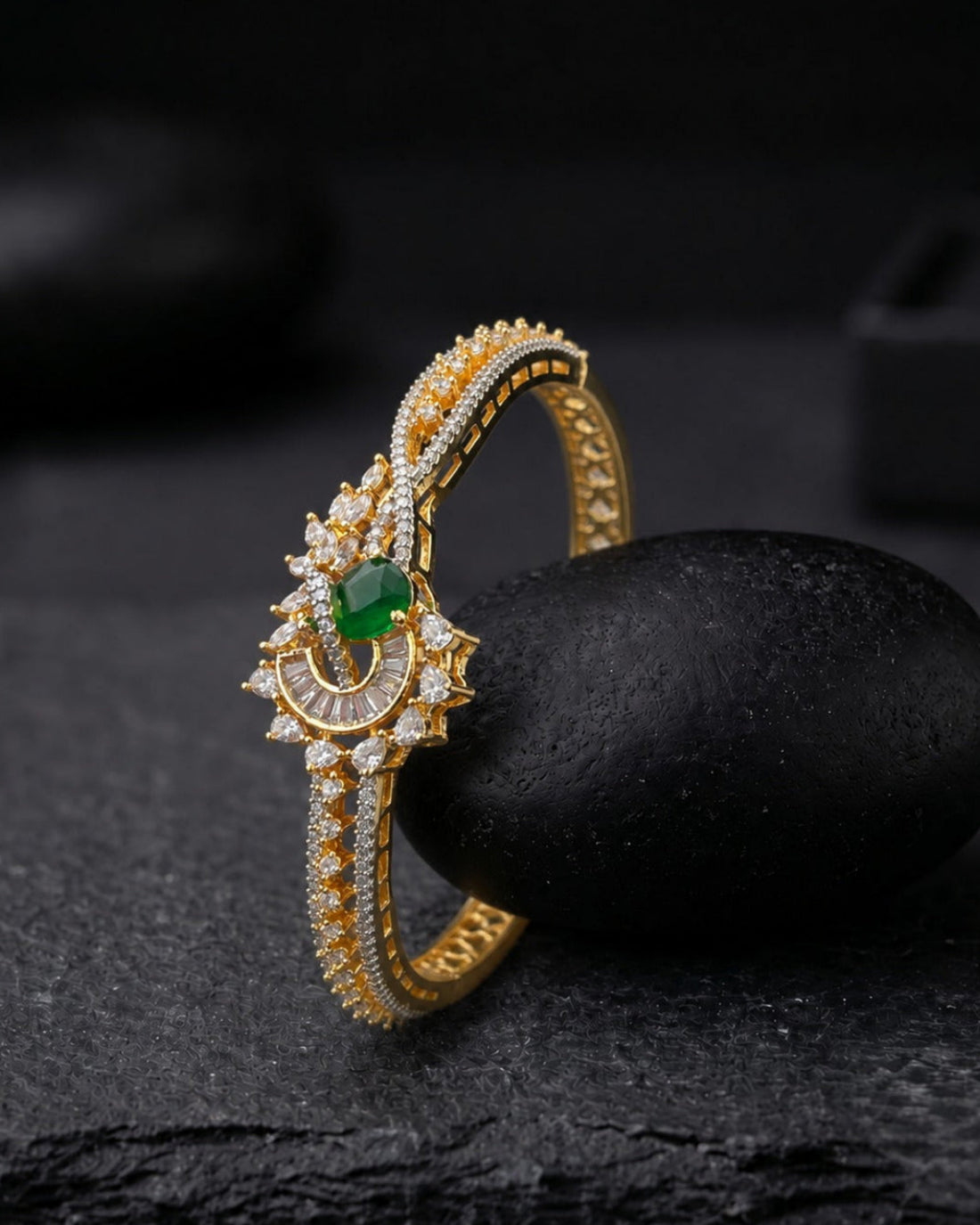 An ornate gold ring with a crescent moon design, set with small clear stones and a central green gemstone, displayed on a black stone against a dark background.