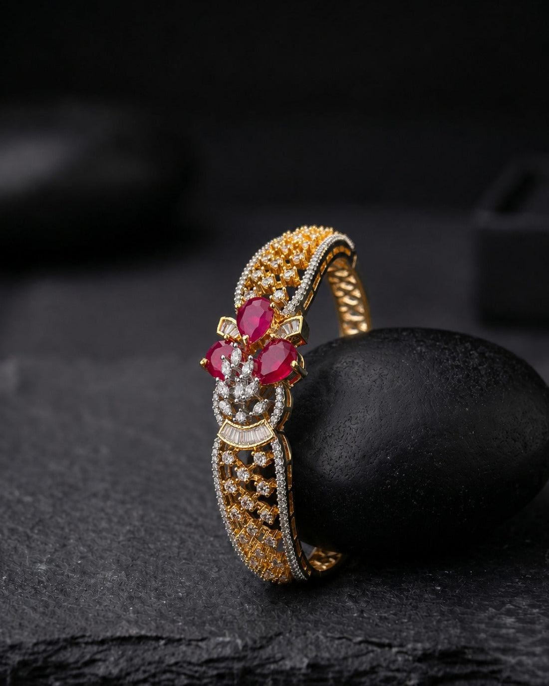 An ornate gold ring with ruby gemstones and small clear stones, displayed on a dark stone against a dark background.