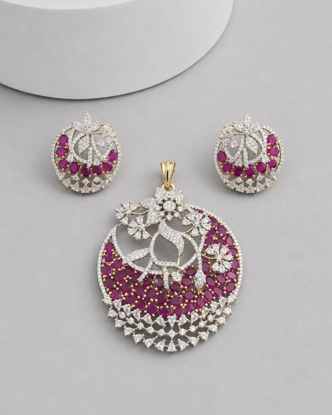 An ornate jewelry set featuring a large crescent-shaped pendant, a matching pair of round earrings, and a small gold clasp, all decorated with white stones and deep pink gemstones on a light gray background.