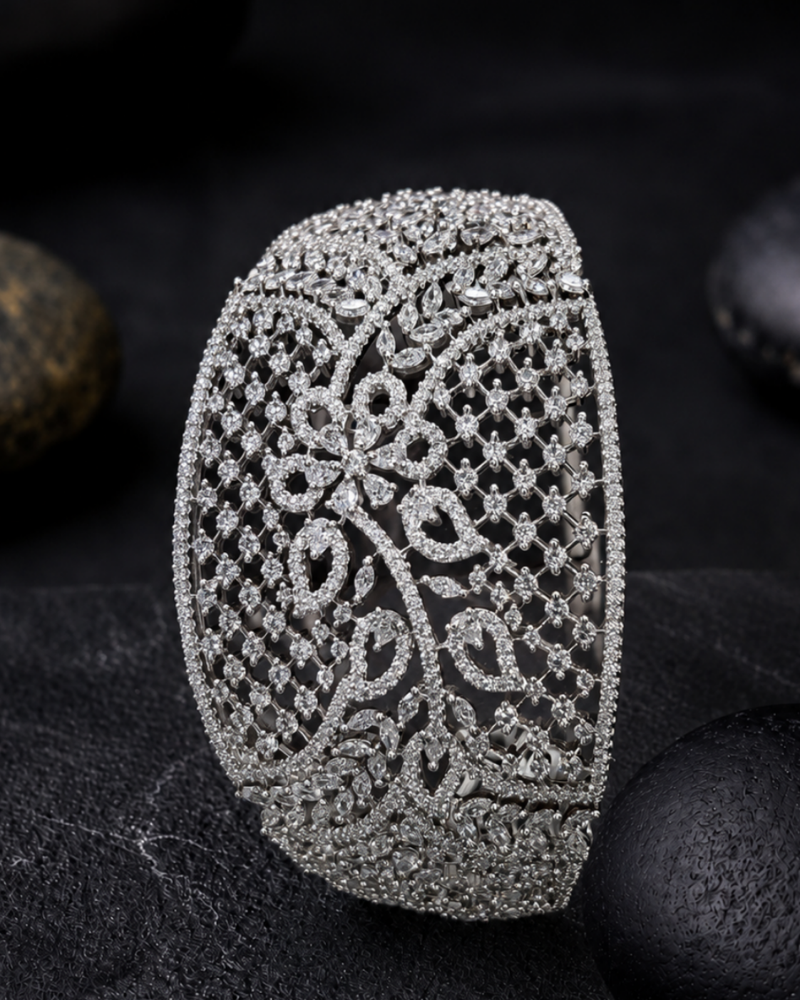 An ornate silver-toned cuff bracelet encrusted with clear stones, featuring a detailed floral filigree pattern, displayed on a dark textured surface with smooth black stones in the background.