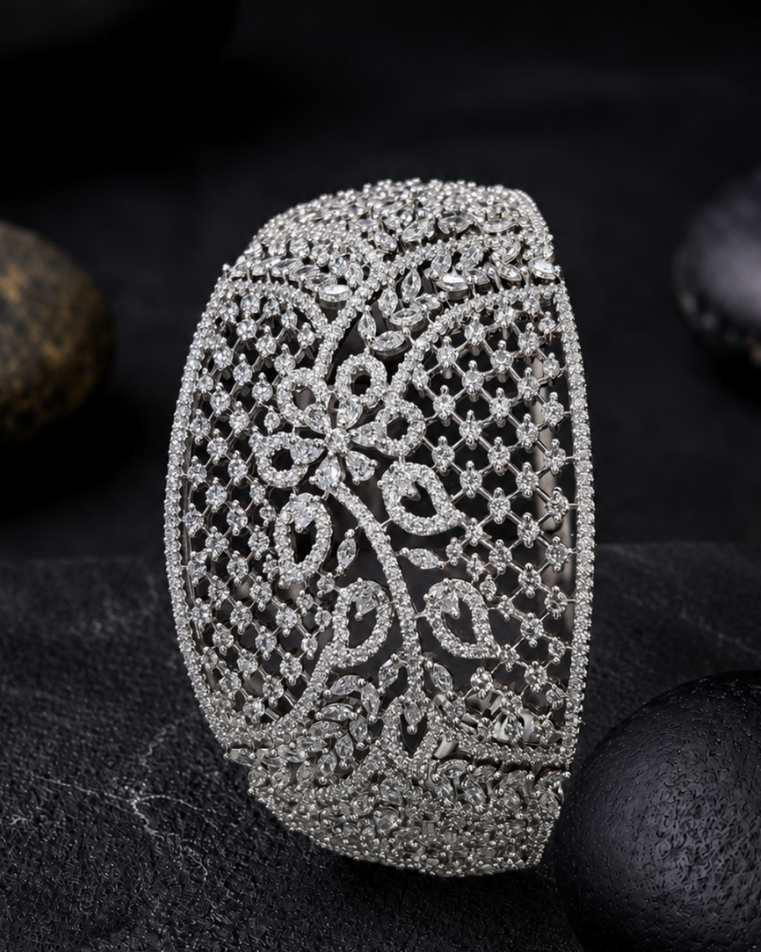An ornate silver-toned cuff bracelet encrusted with clear stones, featuring a detailed floral filigree pattern, displayed on a dark textured surface with smooth black stones in the background.