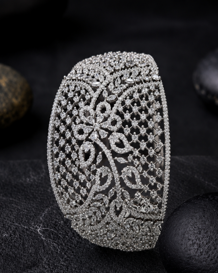 An ornate silver-toned cuff bracelet encrusted with clear stones, featuring a detailed floral filigree pattern, displayed on a dark textured surface with smooth black stones in the background.