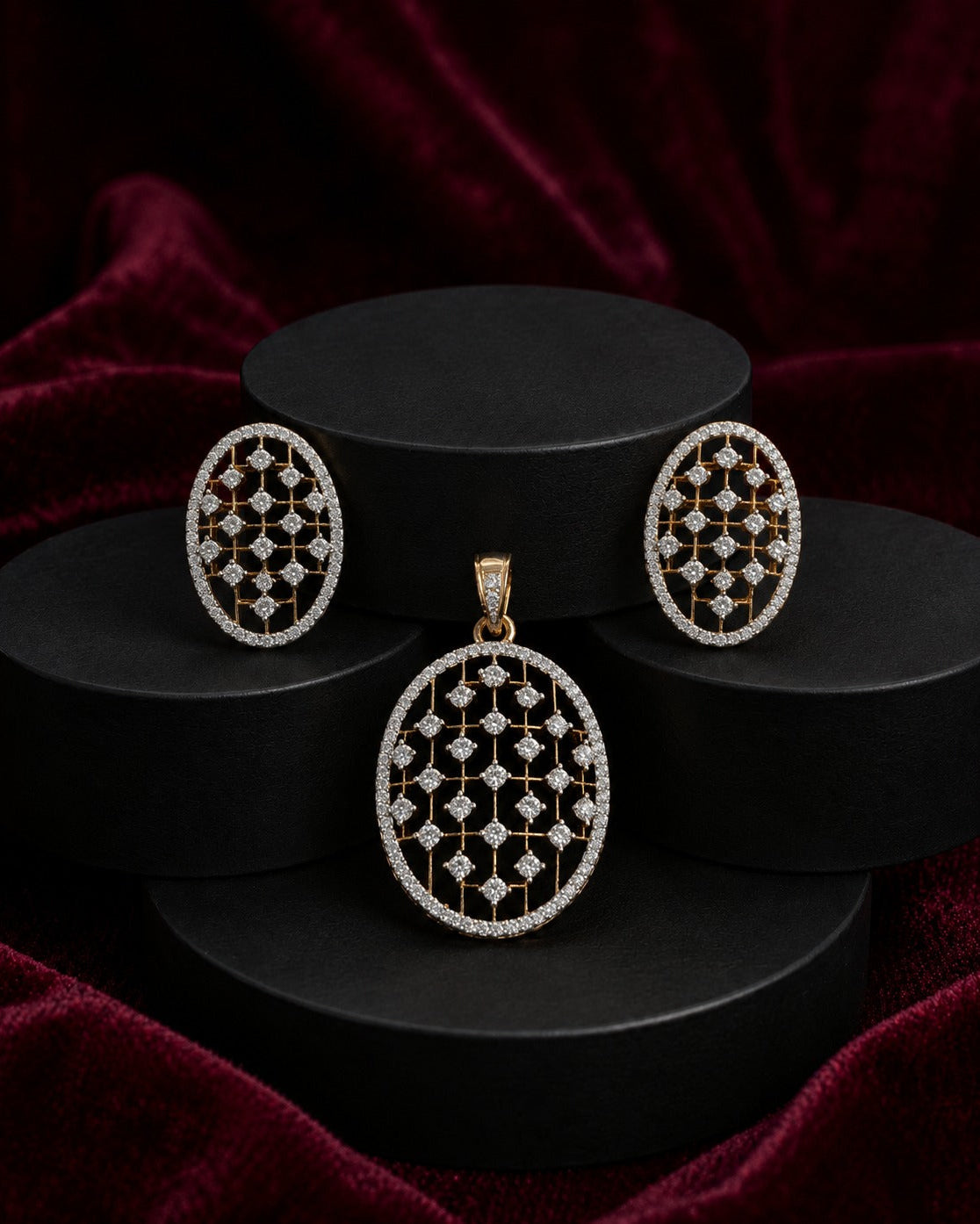 An oval jewelry set featuring a pendant and matching earrings with an intricate openwork geometric pattern in gold and clear stones, displayed on black cylindrical pedestals against a dark red velvet background.