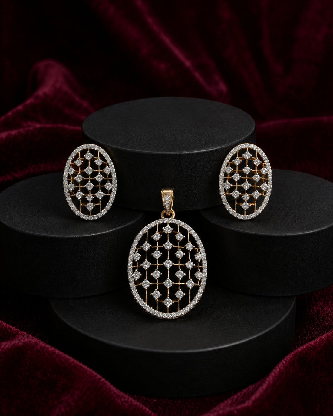 An oval jewelry set featuring a pendant and matching earrings with an intricate openwork geometric pattern in gold and clear stones, displayed on black cylindrical pedestals against a dark red velvet background.