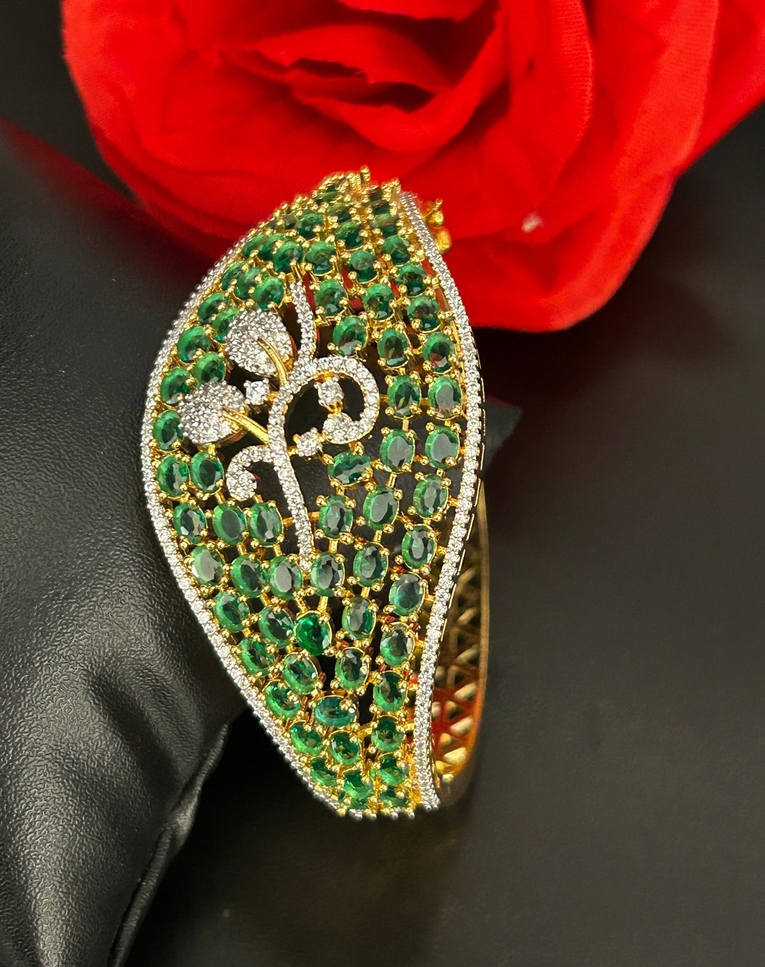 Decorative brooch with green gemstones and gold accents on a black surface with a red flower in the background.