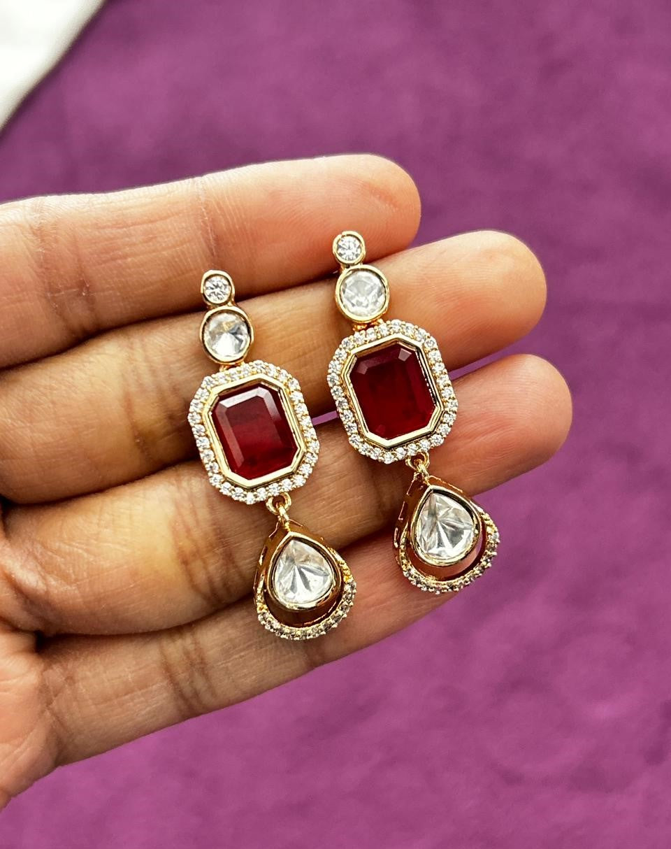 Decorative earrings with red and clear gemstones held in a hand against a purple background