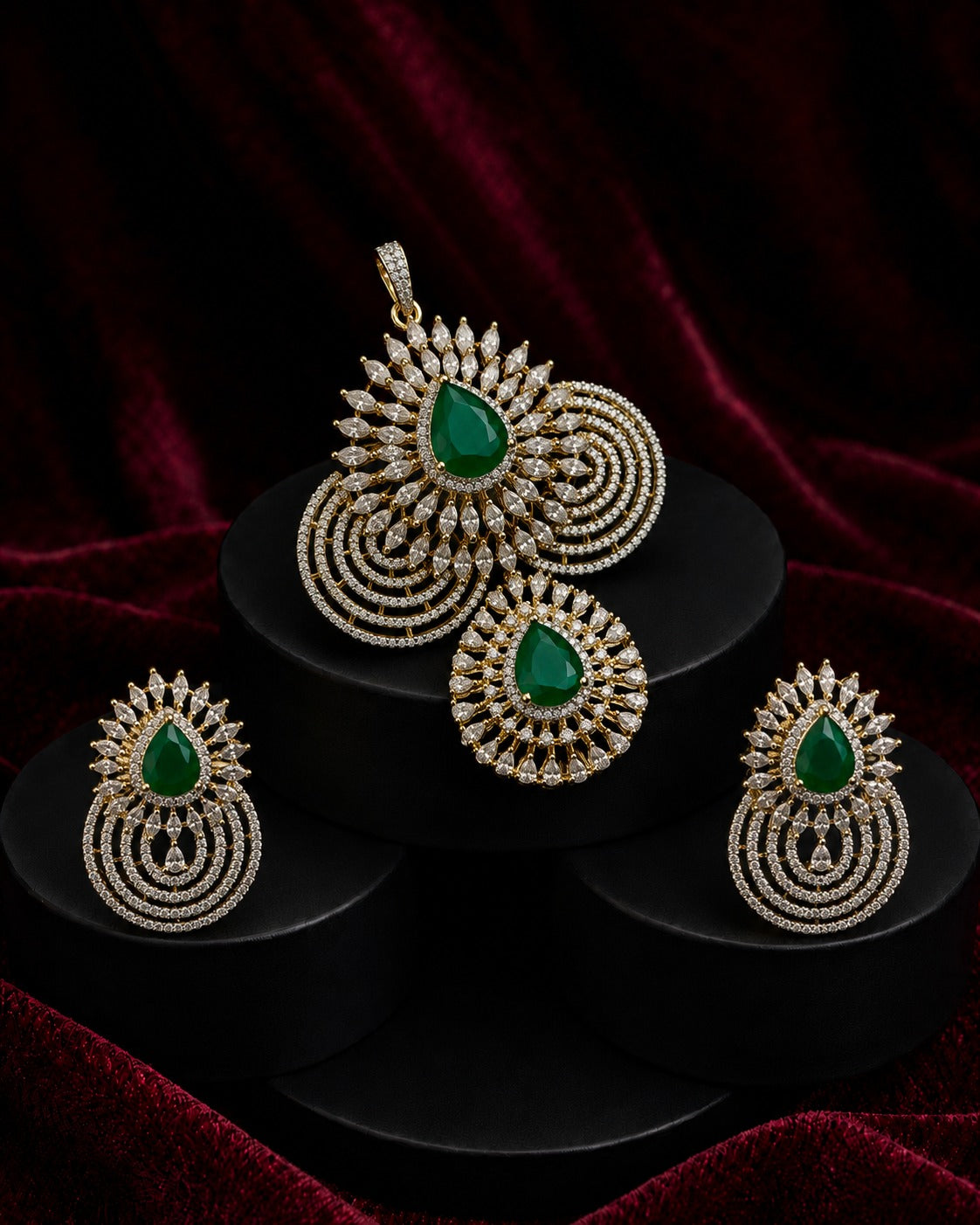 Gold-toned traditional Indian jewelry set featuring a pendant, earrings, and a matching headpiece, each adorned with a green teardrop stone and intricate gold filigree, displayed on black stands against a dark red velvet background.