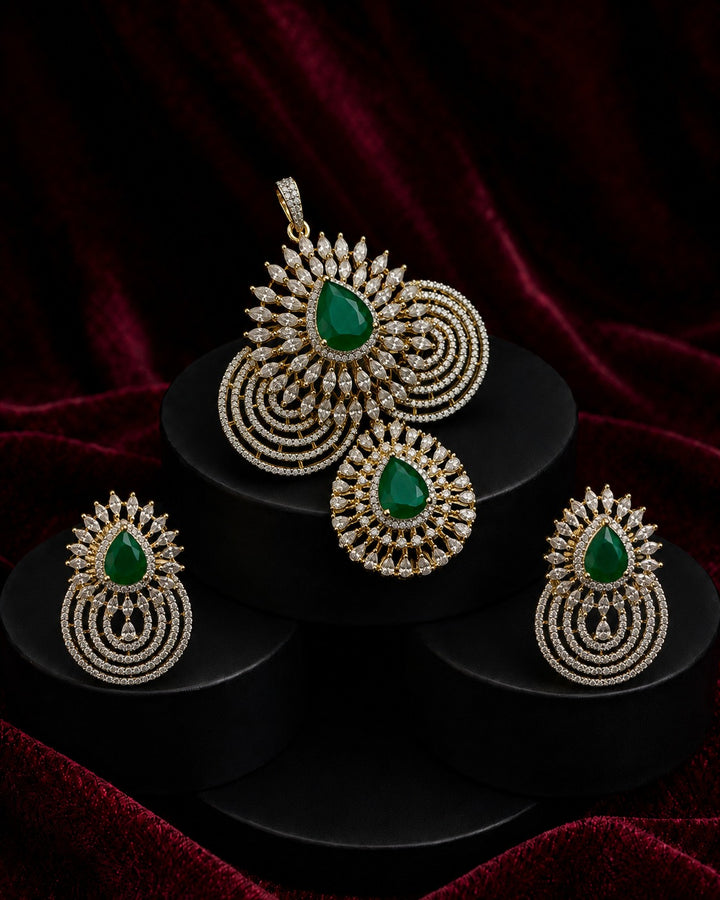 Gold-toned traditional Indian jewelry set featuring a pendant, earrings, and a matching headpiece, each adorned with a green teardrop stone and intricate gold filigree, displayed on black stands against a dark red velvet background.