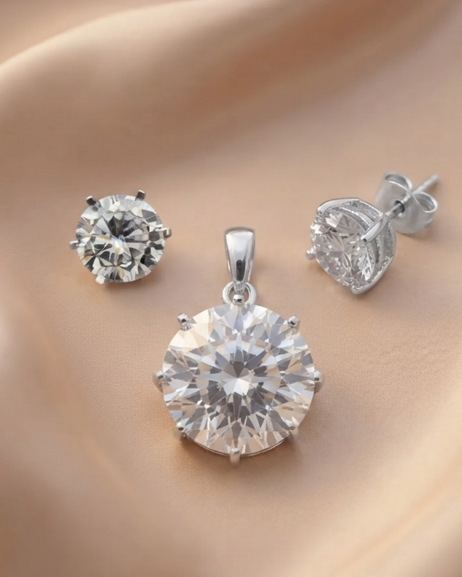 A silver jewelry set featuring a round-cut clear gemstone pendant, a matching round stud earring, and a heart-shaped clear gemstone stud earring with a butterfly back, displayed on a draped beige satin fabric.