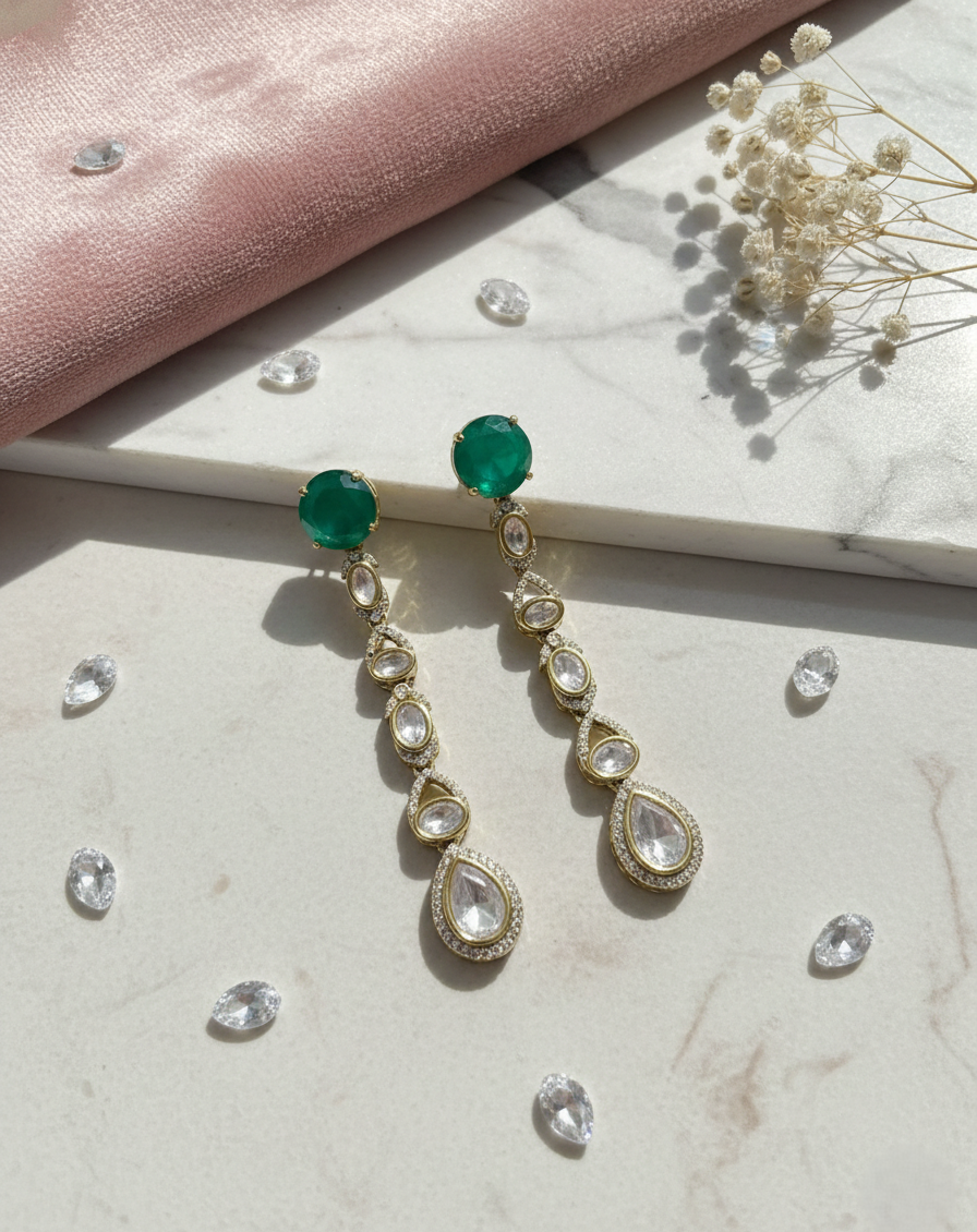 Gold earrings with green and clear gemstones on a marble surface with pink fabric and dried flowers.