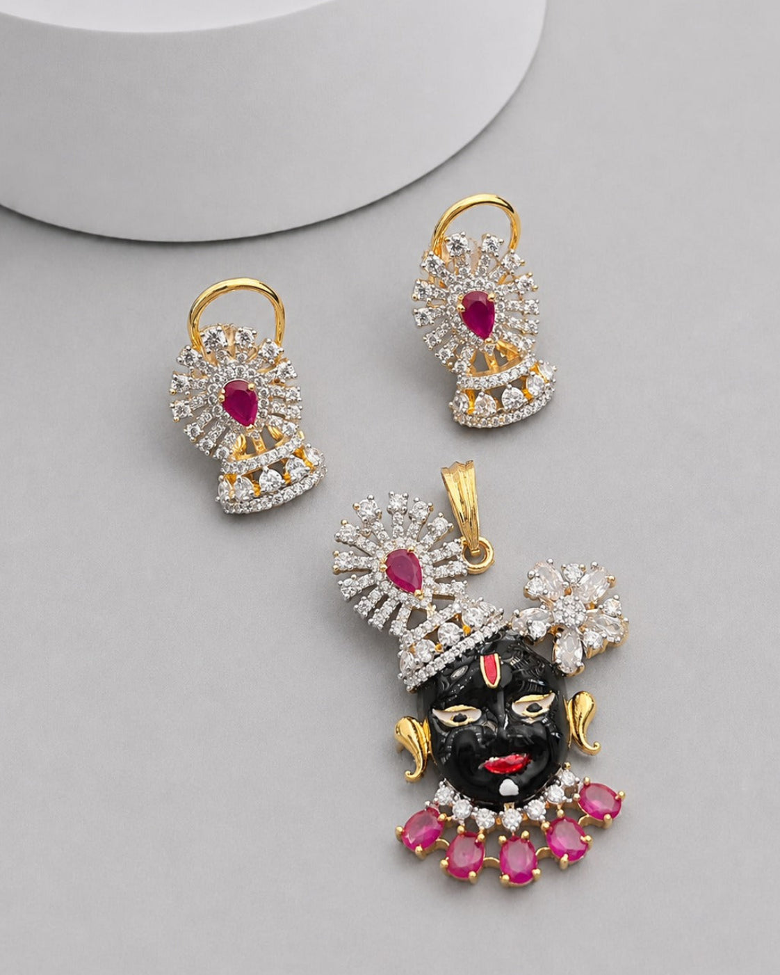 Gold-plated jewelry set featuring a black Lord Krishna pendant and matching earrings, embellished with clear crystals and pink gemstones, displayed on a light gray background.