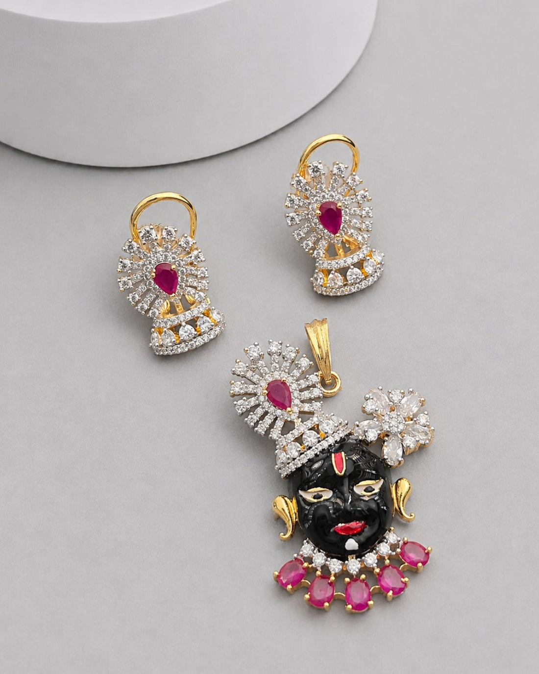 Gold-plated jewelry set featuring a black Lord Krishna pendant and matching earrings, embellished with clear crystals and pink gemstones, displayed on a light gray background.