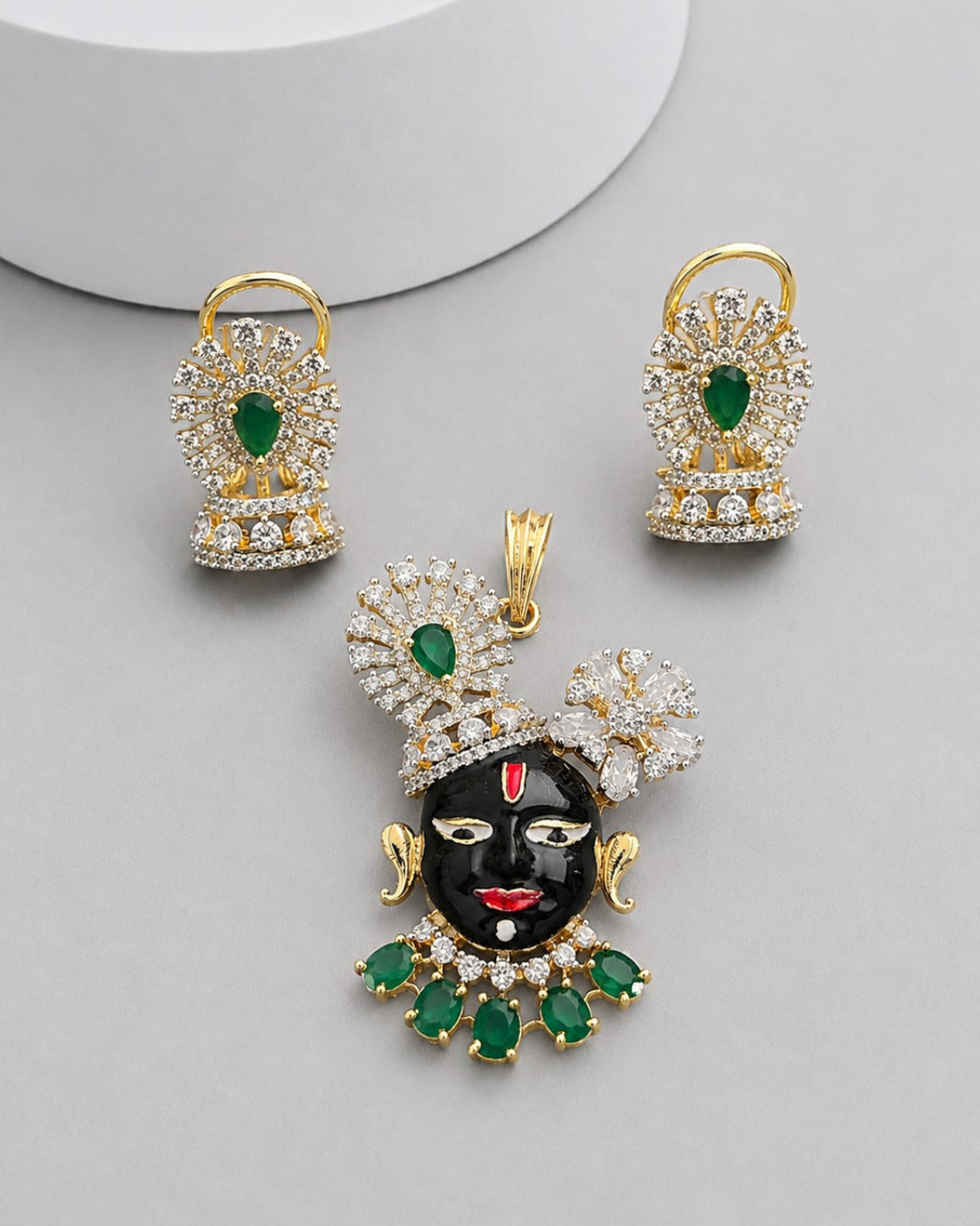 Gold-plated jewelry set featuring a black Lord Krishna pendant and matching earrings, embellished with green emerald-cut stones and clear cubic zirconia crystals, displayed on a light gray background.