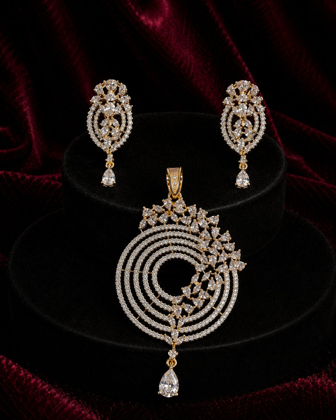 Gold-plated jewelry set featuring a large circular pendant, a matching pair of drop earrings, and a teardrop-shaped drop, all embellished with clear stones and set against a dark velvet background.