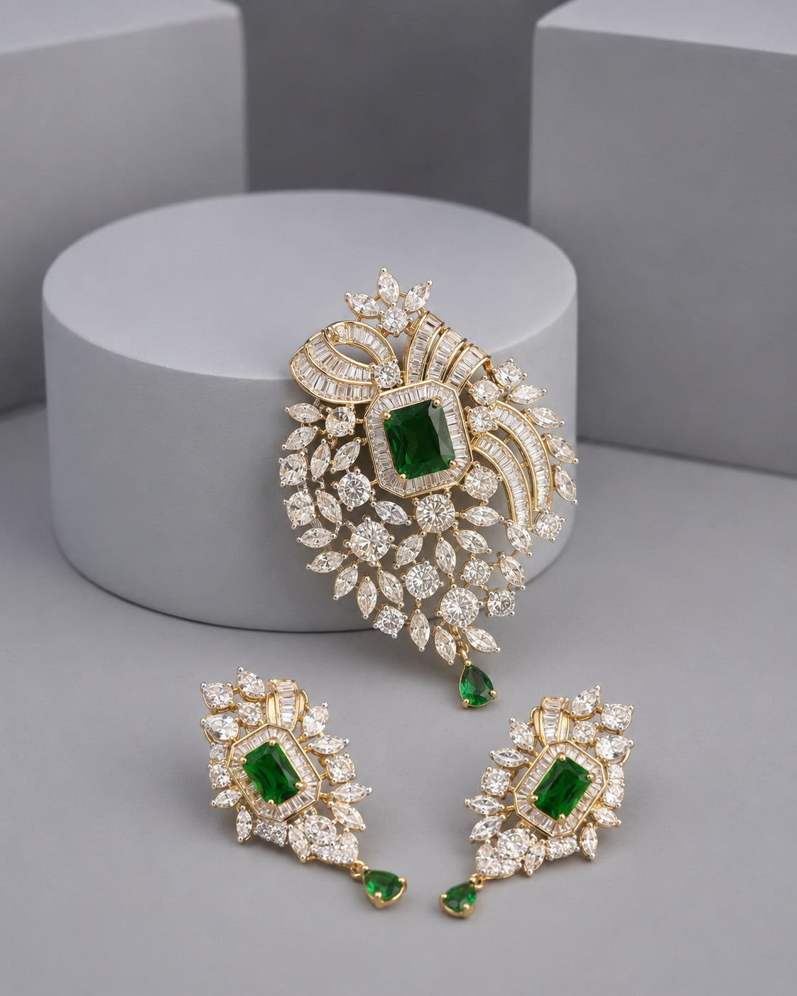 Gold-plated jewelry set featuring a large embroidered brooch and matching drop earrings, embellished with clear crystals and green emerald-cut stones, displayed on a gray studio background.