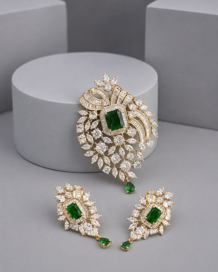 Gold-plated jewelry set featuring a large embroidered brooch and matching drop earrings, embellished with clear crystals and green emerald-cut stones, displayed on a gray studio background.