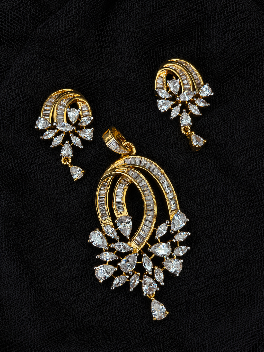 Gold-tone jewelry set featuring a large pendant and matching earrings, both adorned with clear gemstones, displayed on a textured black fabric background.