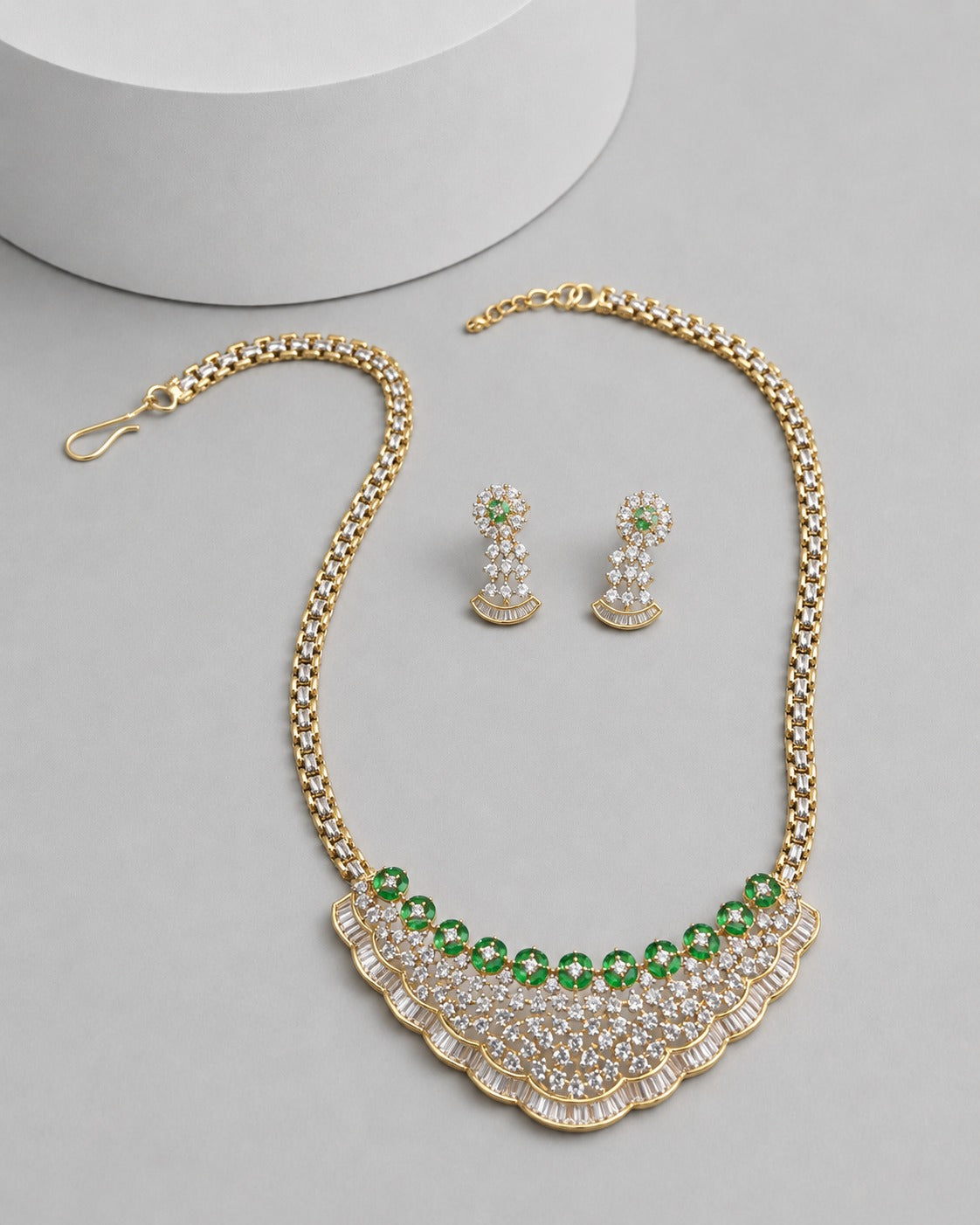 Gold-tone necklace and matching drop earrings with green faceted stones and clear crystal-like accents, arranged on a light gray surface with a white cylindrical prop.