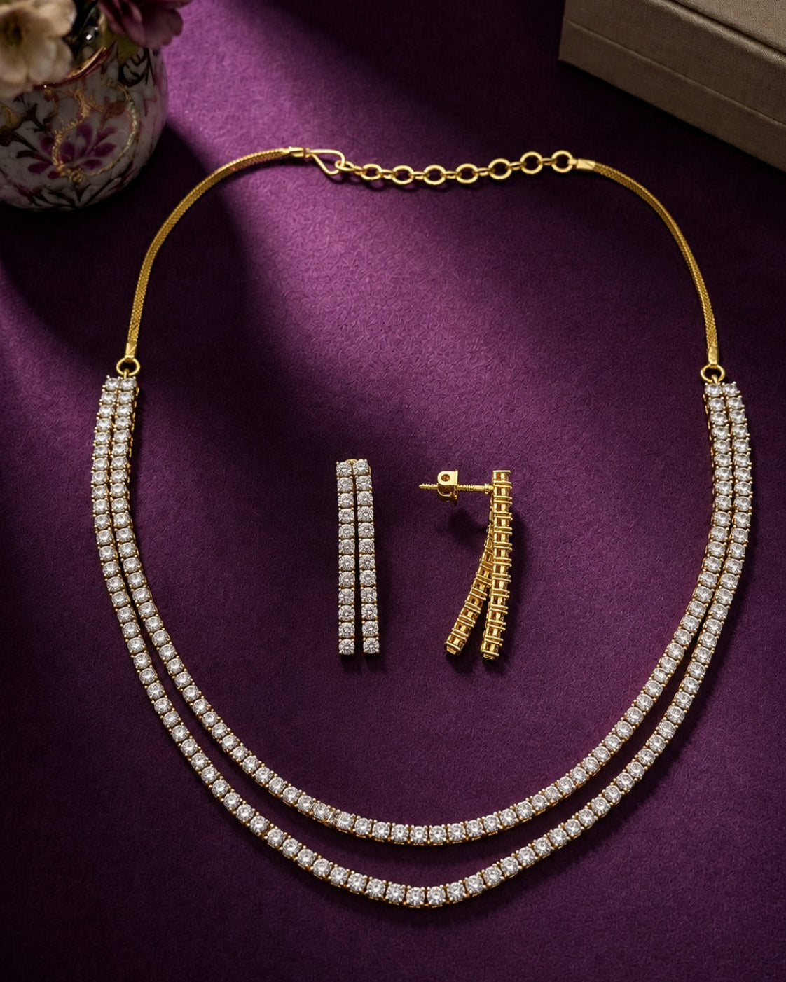 Gold-tone necklace and matching earrings set with clear stones, arranged on a purple satin-like surface with decorative props in the corners.