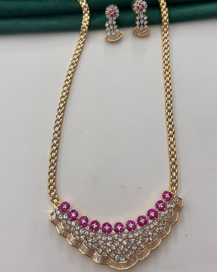 Gold-tone necklace and matching earrings set with pink gemstone accents and clear rhinestone detailing, displayed on a white surface with a dark green fabric backdrop.